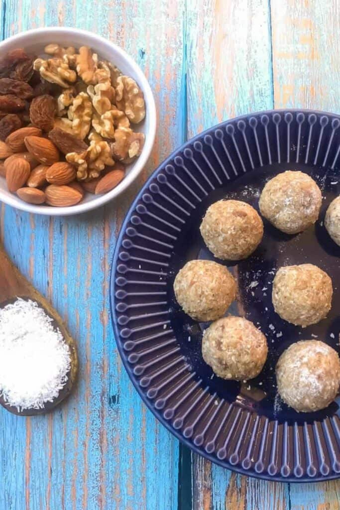 Plate of 10 minute energy balls arranged on a dark blue dish, coated lightly in shredded coconut with a soft, textured surface. Nearby bowls of almonds, walnuts, raisins, and extra coconut.
