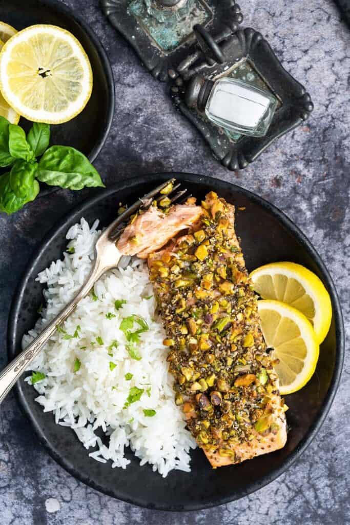 Flaky salmon coated with a crunchy pistachio and herb crust, served with fluffy white rice and fresh lemon slices on a black plate. This 15-minute pistachio crusted salmon is a quick, flavorful dinner with a crisp nutty topping and bright citrus flavor.