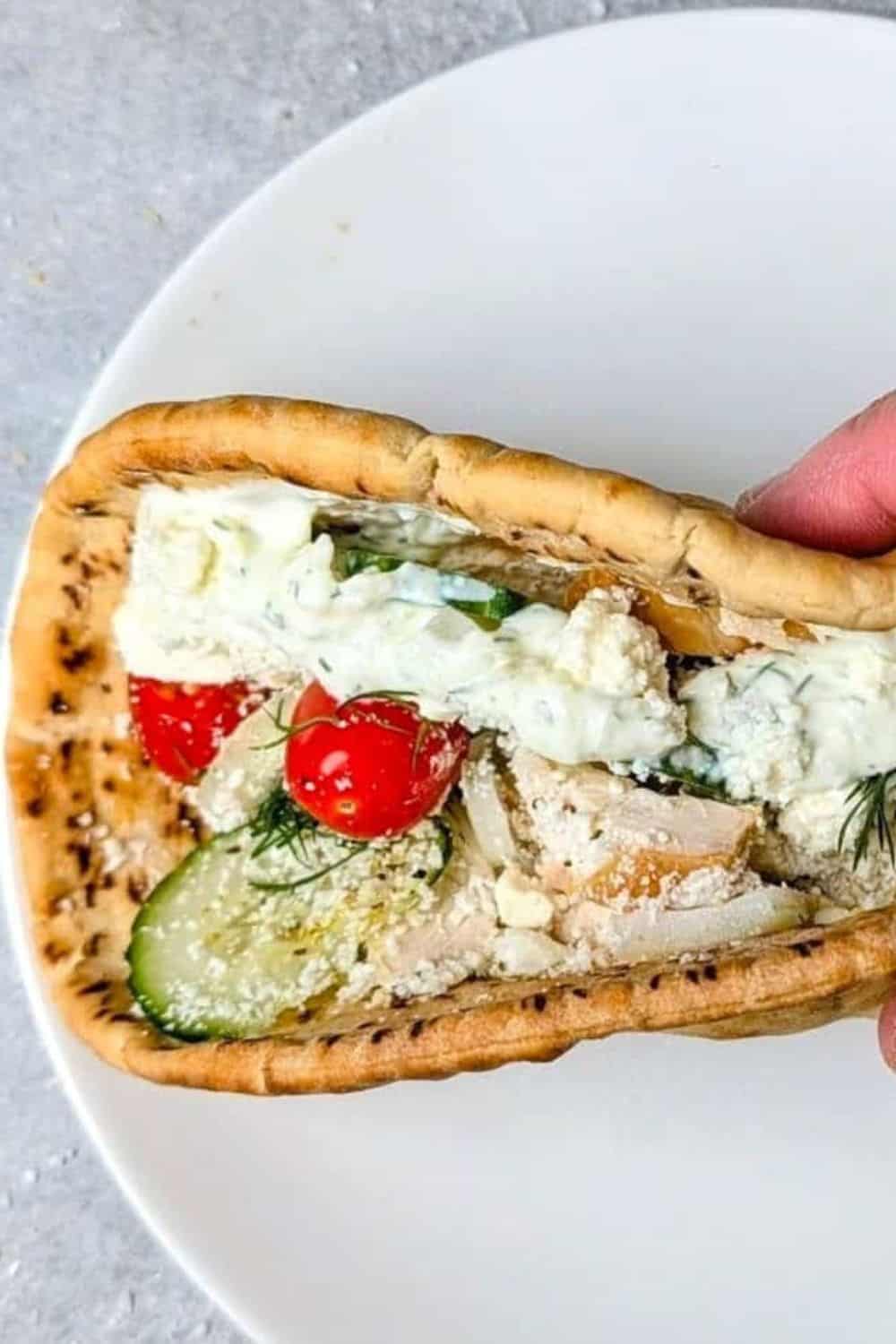 Warm pita filled with sliced chicken, creamy tzatziki, cucumber, cherry tomatoes, fresh dill, and crumbled feta, held over a white plate. These 20 minute gyros are a fresh and easy weeknight meal with classic Mediterranean flavors.