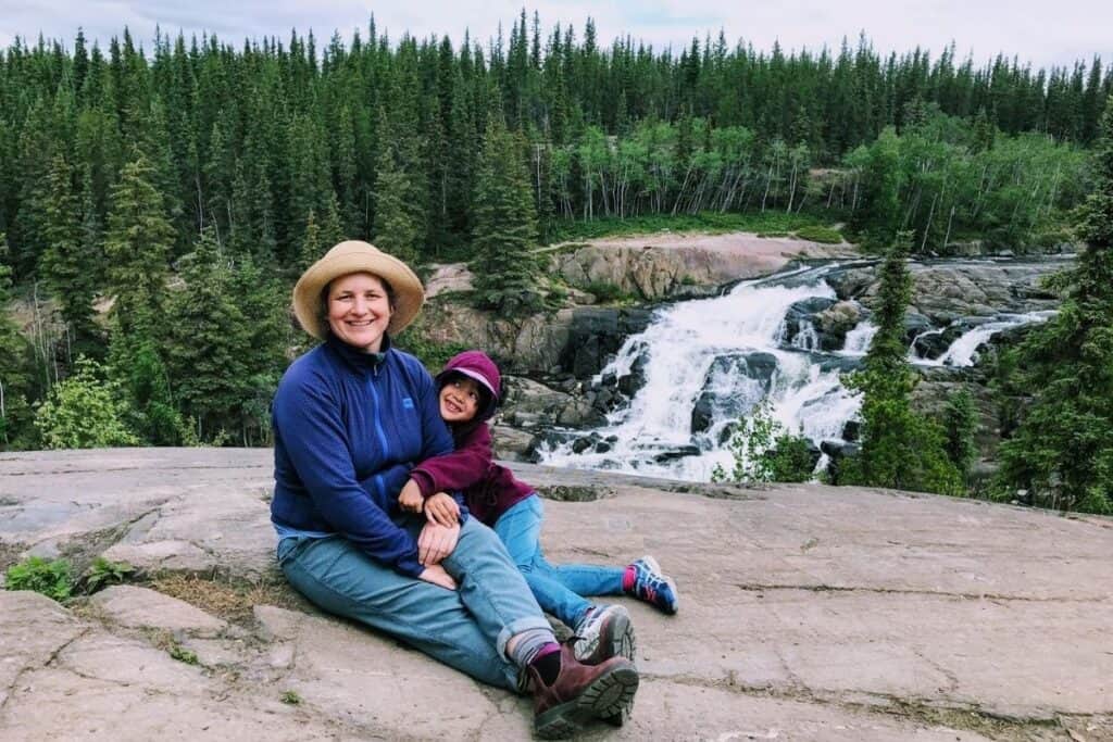 Hiking near Yellowknife at Cameron Falls view point, Hidden Lake Territorial Park. Mother and daughter sitting on rock, waterfall and forest in background.