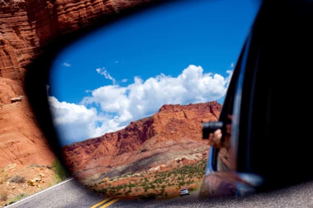 How to take a cheap Family Road Trip. Driving through local national park with red rocks, taking a photo out the window, captured in side mirror.