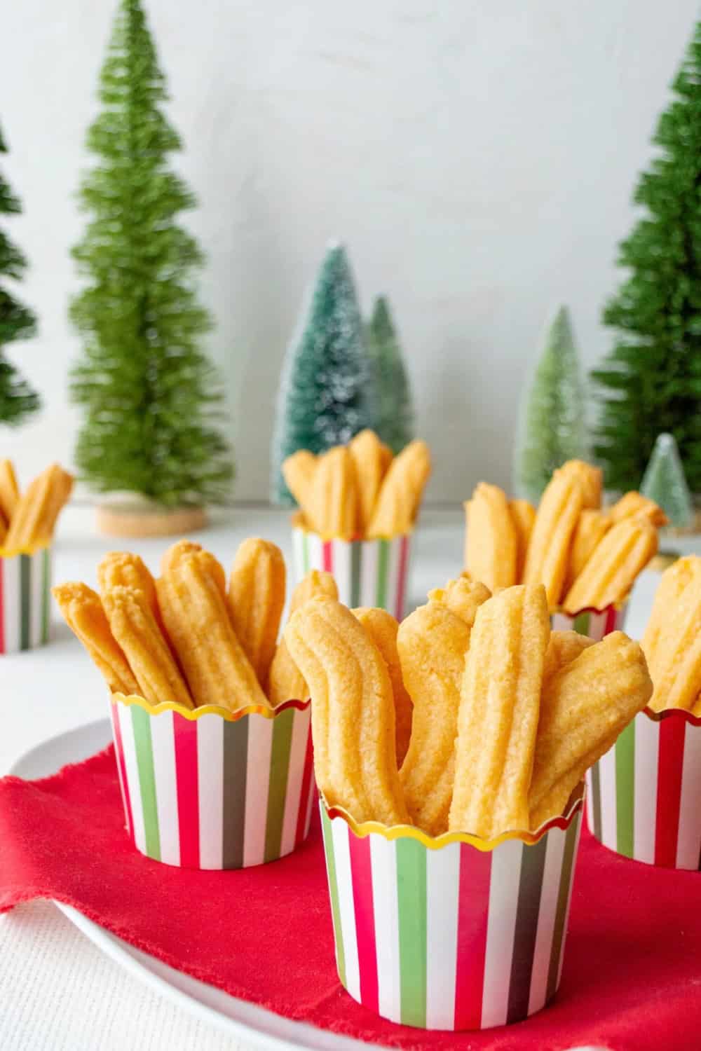 Golden Southern cheese straws served upright in red green and white striped cups. The cheese straws are arranged on a red cloth with miniature Christmas trees in the background.