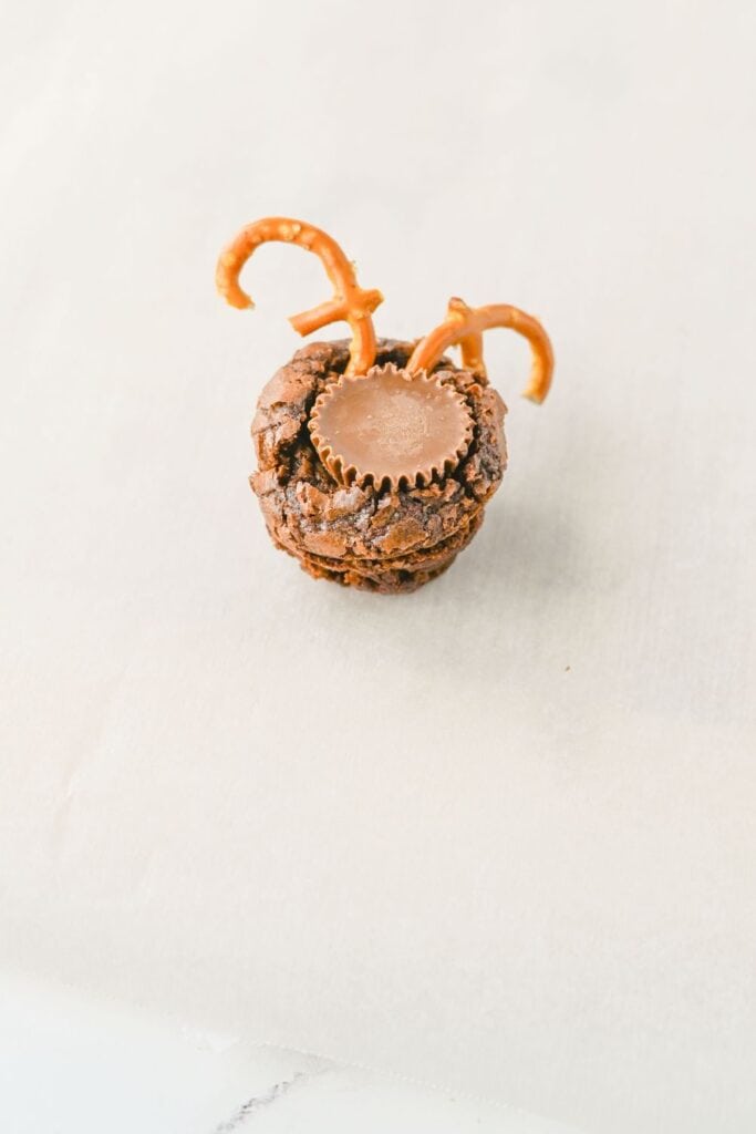 Partially decorated reindeer brownie with a Reese’s mini cup pressed into the center and two pretzel pieces forming antlers on top. The treat is set on a parchment-lined surface, mid-assembly.