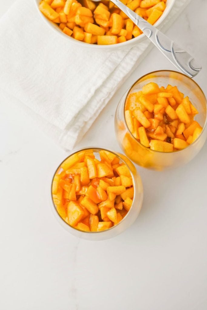 Overhead view of two glass cups filled with a layer of spiced, caramel-coated diced apples. A white bowl of the same apple mixture with a spoon rests nearby on a folded white towel, showing the step-by-step dessert assembly.