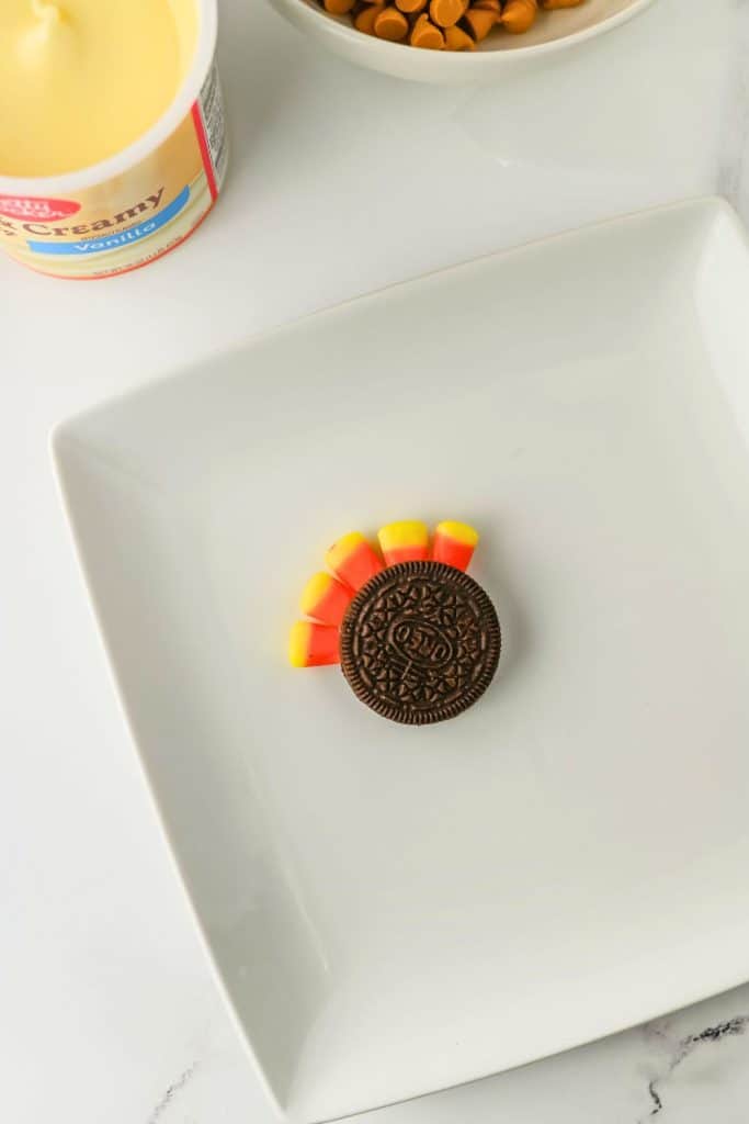 Oreo cookie on a white plate with five pieces of candy corn inserted along one side to resemble tail feathers, beginning the assembly of an turkey Oreo cookies. Vanilla frosting and a bowl of butterscotch chips are visible in the background, showing ingredients used for decorating.