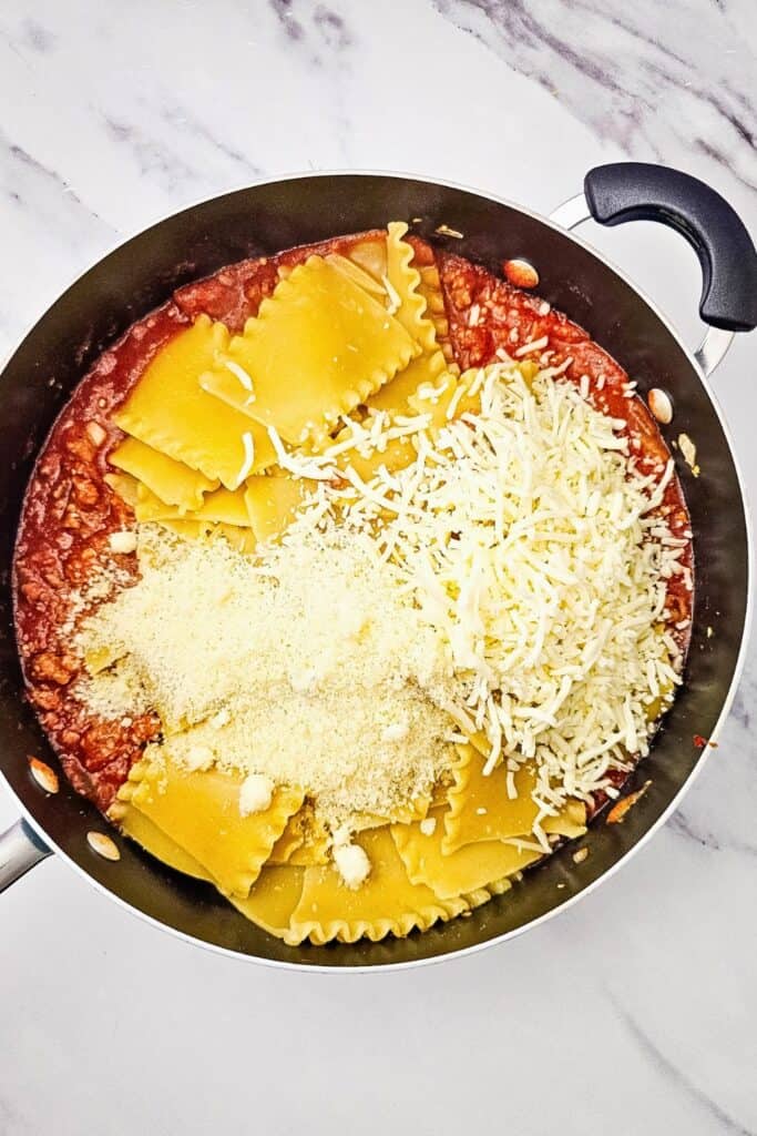 Cooked lasagna noodles and Italian sausage meat sauce topped with shredded mozzarella and grated Parmesan. The cheeses are ready to be mixed with the noodles and sauce for the sheet pan lasagna.
