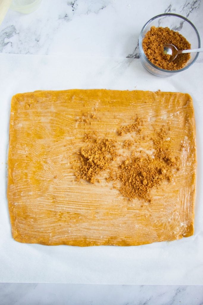 Rectangular sheet of rolled-out pumpkin cookie dough brushed with melted butter and partially topped with a brown sugar and cinnamon mixture. A bowl of the cinnamon sugar filling with a spoon sits nearby on the marble surface.