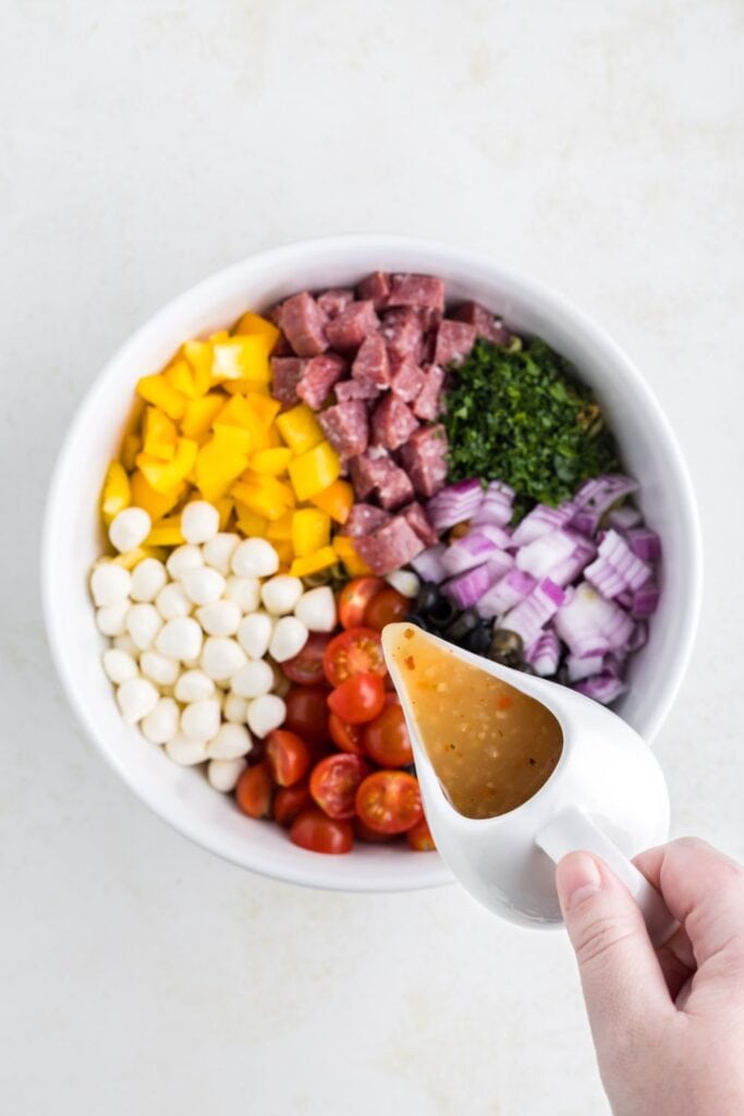 A hand pouring Italian dressing from a cup into a large white ceramic bowl filled with sections of cut up ingredients for zesty Italian pasta salad including cherry tomatoes, yellow peppers, parsley, salami, red onion, black olives, mini mozzarella balls.