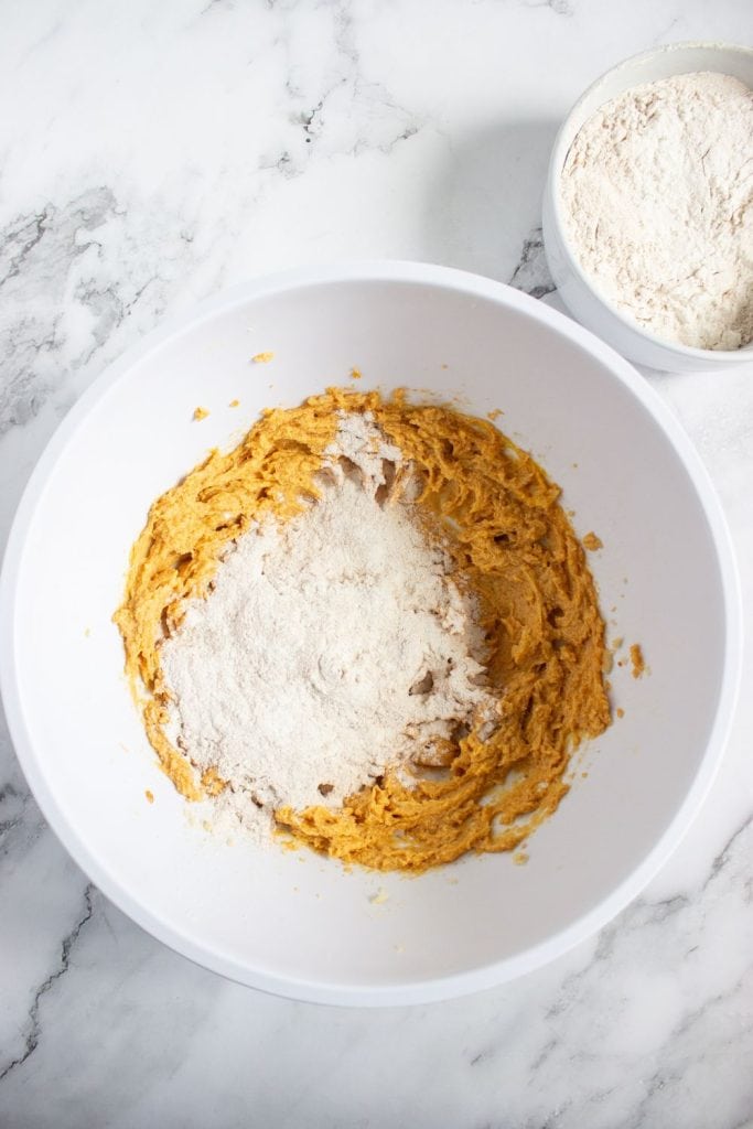 Dry flour mixture being added to a bowl of wet pumpkin cookie dough, midway through combining the ingredients. The mixing bowl rests on a marble surface, with the remaining dry mix visible in a separate bowl nearby.