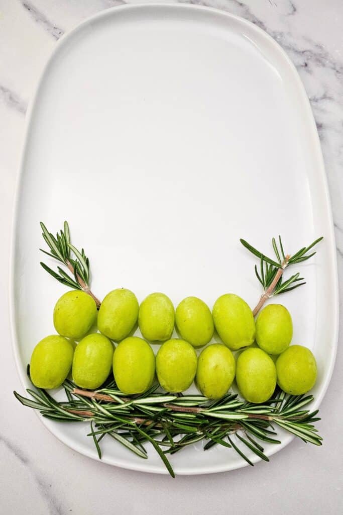 Two rows of fresh green grapes arranged over rosemary sprigs on a white oval platter, forming the bottom layer of an easy Christmas tree fruit tray.