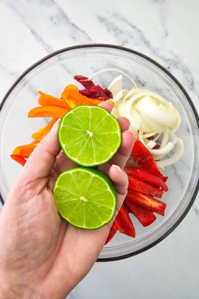 Two fresh lime halves are held over a glass bowl filled with raw steak. Sliced red and orange bell peppers and white onions are ready to be tossed with the lime juice and fajita seasoning.