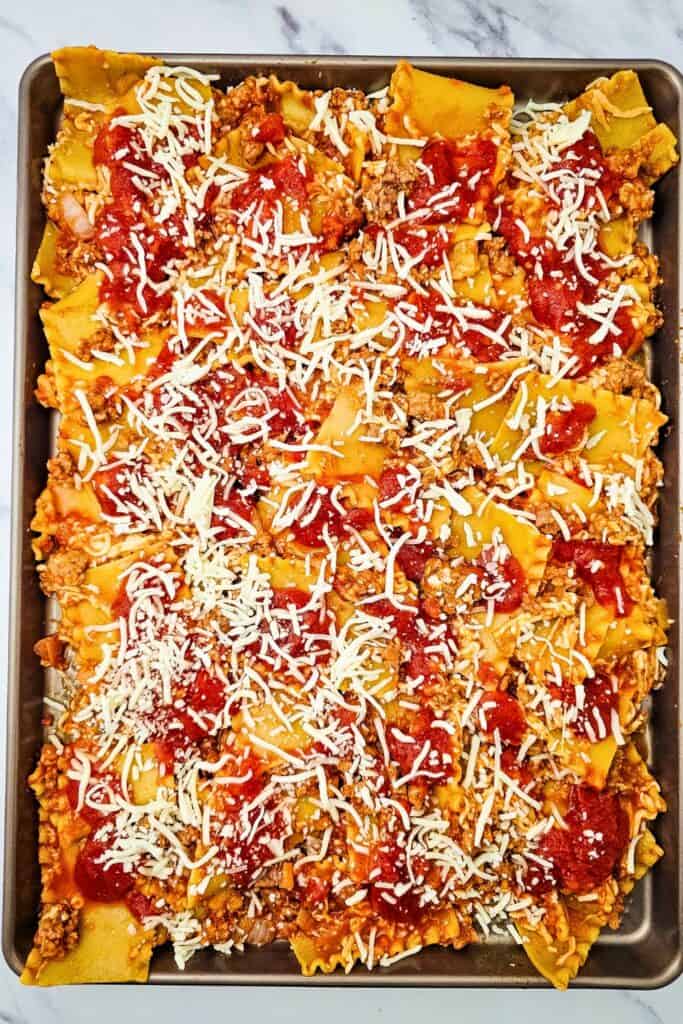 Sheet pan lasagna topped with tomato pasta sauce and shredded mozzarella over broken noodles and Italian sausage. The cheese is scattered evenly across the pan before baking.