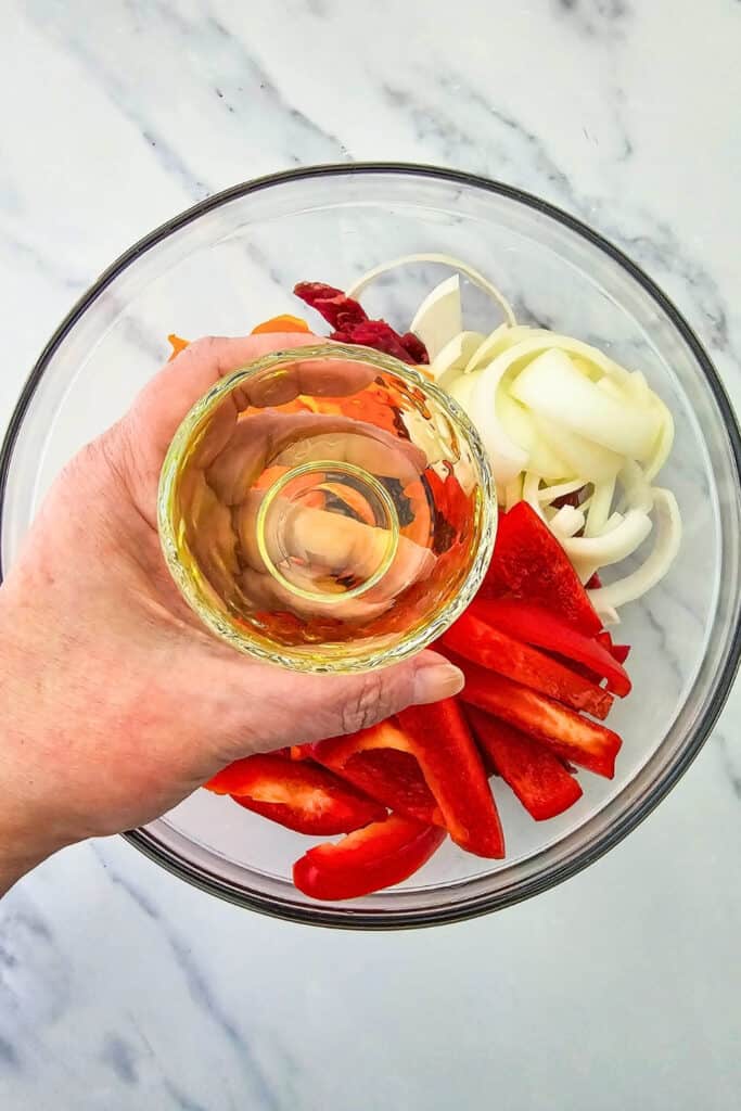 A small glass bowl of olive oil is held over a large mixing bowl filled with raw steak strips. Sliced red and orange bell peppers and white onions are ready to be coated with the oil for steak fajitas.