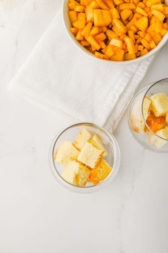 Overhead view of dessert assembly showing two glass cups partially filled with pound cake cubes and caramel sauce. A large bowl of spiced apple filling sits nearby on a white towel, ready for layering in the fall-inspired trifles.