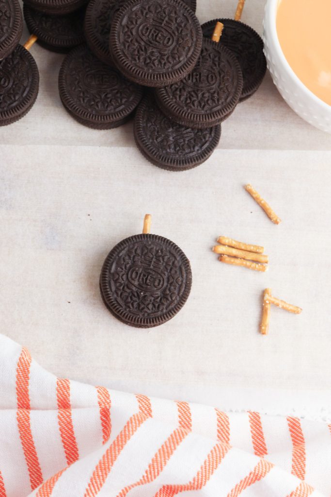 A stack of Oreo cookies sits on parchment paper, with a few cookies individually shown having pretzel sticks inserted at the top. A small bowl of melted orange candy and broken pretzel pieces are nearby, with an orange and white striped towel at the bottom of the frame.