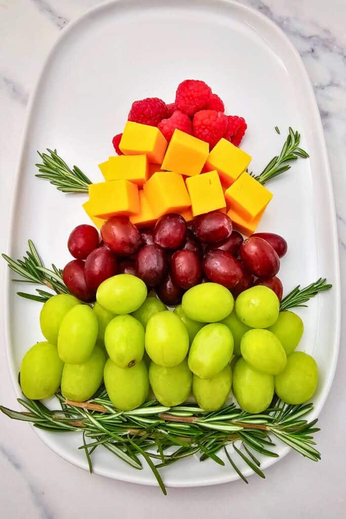 A Christmas tree snack made with layers of green and red grapes, cheddar cheese cubes, and fresh raspberries, with rosemary sprigs forming branches on a white oval serving platter.