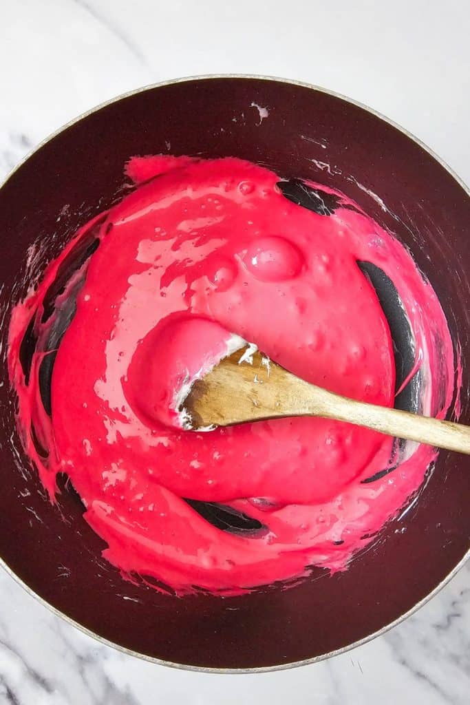 Bright red-pink melted marshmallow mixture in a saucepan, stirred with a wooden spoon to prepare for Halloween rice krispie brains treats.