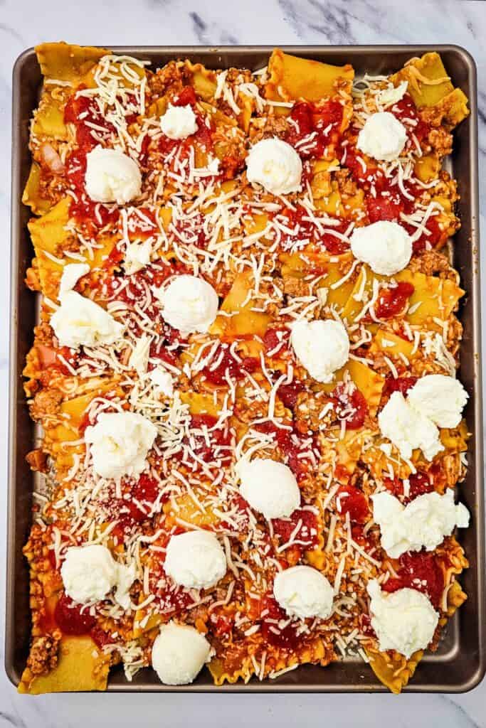 Sheet pan lasagna topped with generous dollops of ricotta cheese over broken noodles with Italian sausage. Tomato sauce and shredded mozzarella are scattered throughout before baking.
