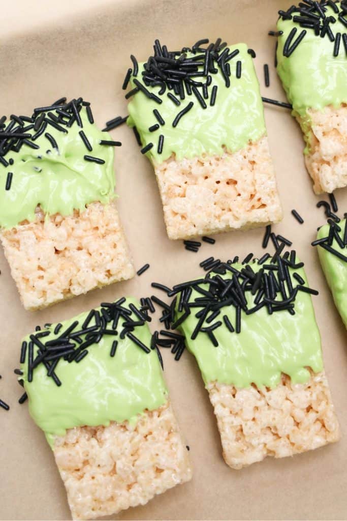 Partially decorated Frankenstein Rice Krispie treats laid out on parchment paper, dipped in green candy coating and topped with black jimmies to resemble hair. The treats are in progress, with candy eyes and facial details yet to be added.