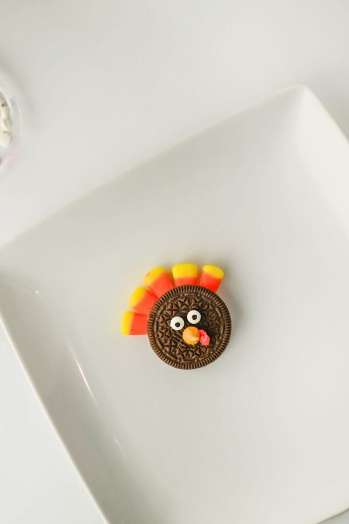 Fully assembled Oreo turkey cookie on a white plate, decorated with candy corn feathers, candy eyes, a butterscotch chip beak, and a red icing wattle.