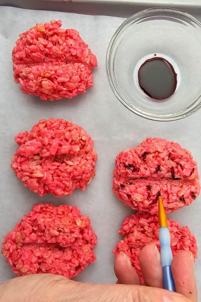 Hand using a small paintbrush to apply dark food coloring into the grooves of bright red rice krispie treats, adding vein-like details to create realistic Halloween brain treats.