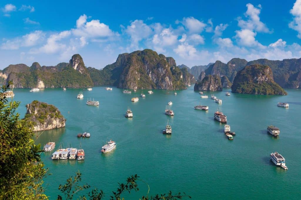 Cruise boats and tour vessels are scattered across the emerald green waters of Ha Long Bay, surrounded by towering limestone islands covered in lush greenery. The scene is framed by blue skies and fluffy white clouds, capturing the natural beauty and scale of this UNESCO World Heritage Site in Vietnam.