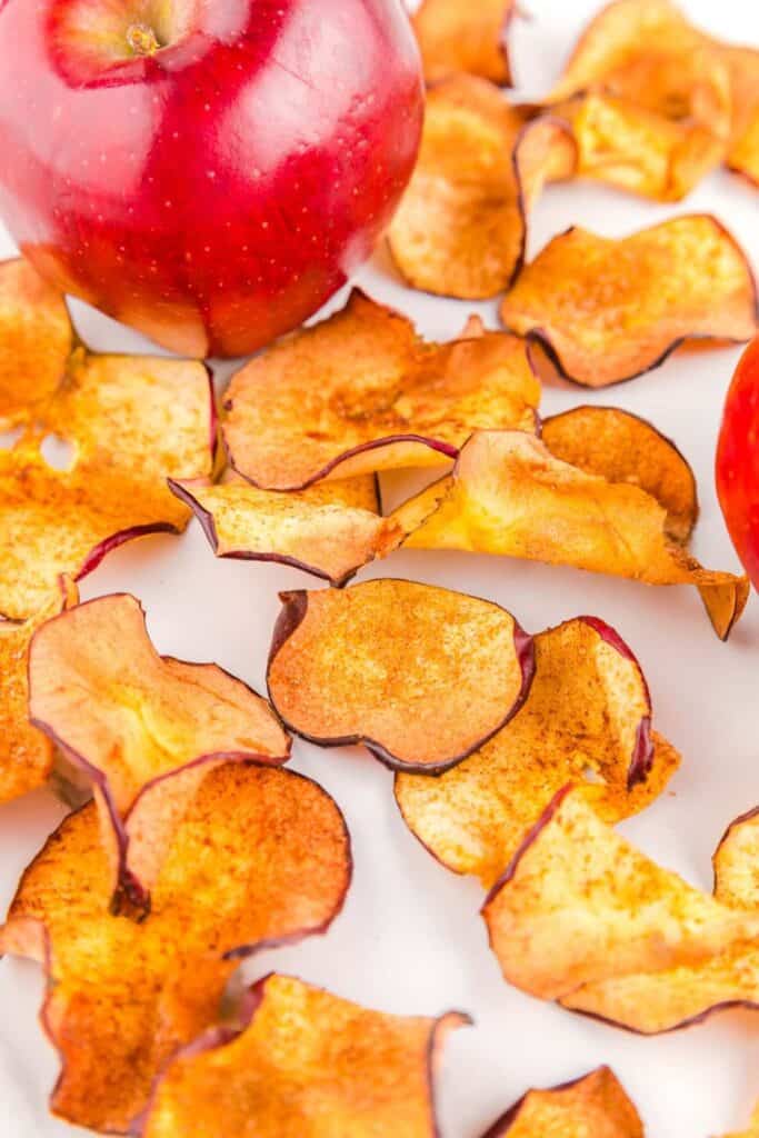 Air fryer Apple chips are spread out on a counter top, with a red apple beside them.