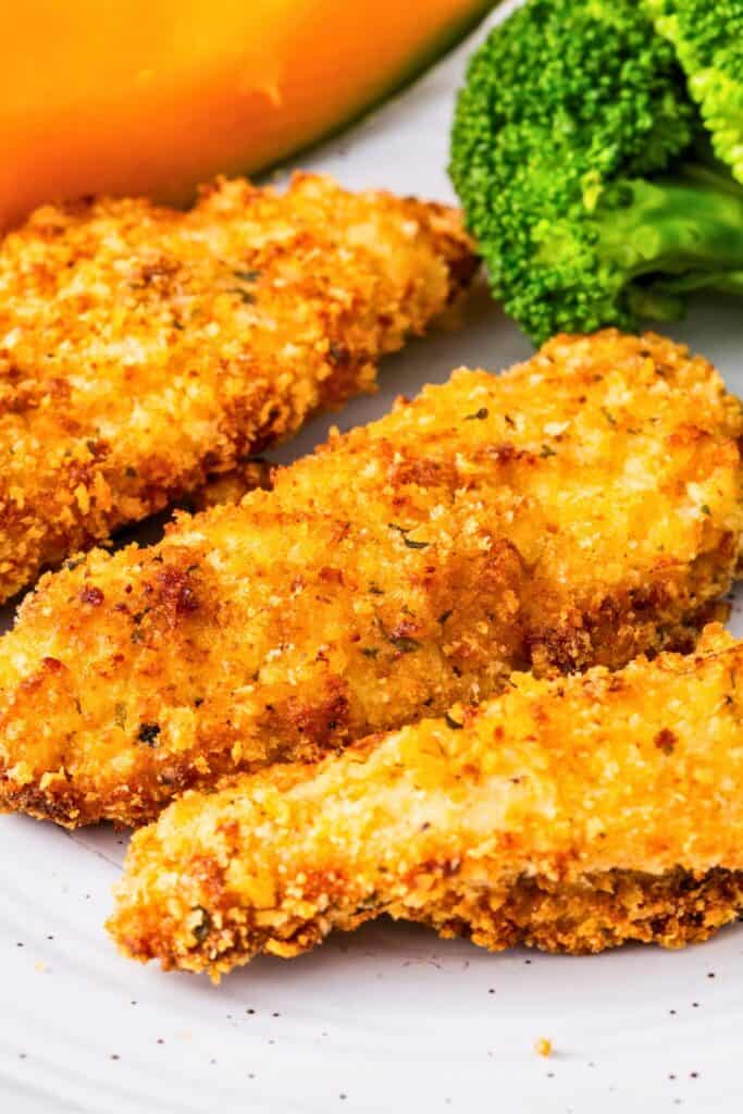 Crispy air fryer chicken strips for kids served on a white plate, coated in a golden brown breadcrumb crust with a crunchy texture. One strip is sliced open to show the tender white chicken inside, with broccoli and a slice of squash in the background for a balanced meal.