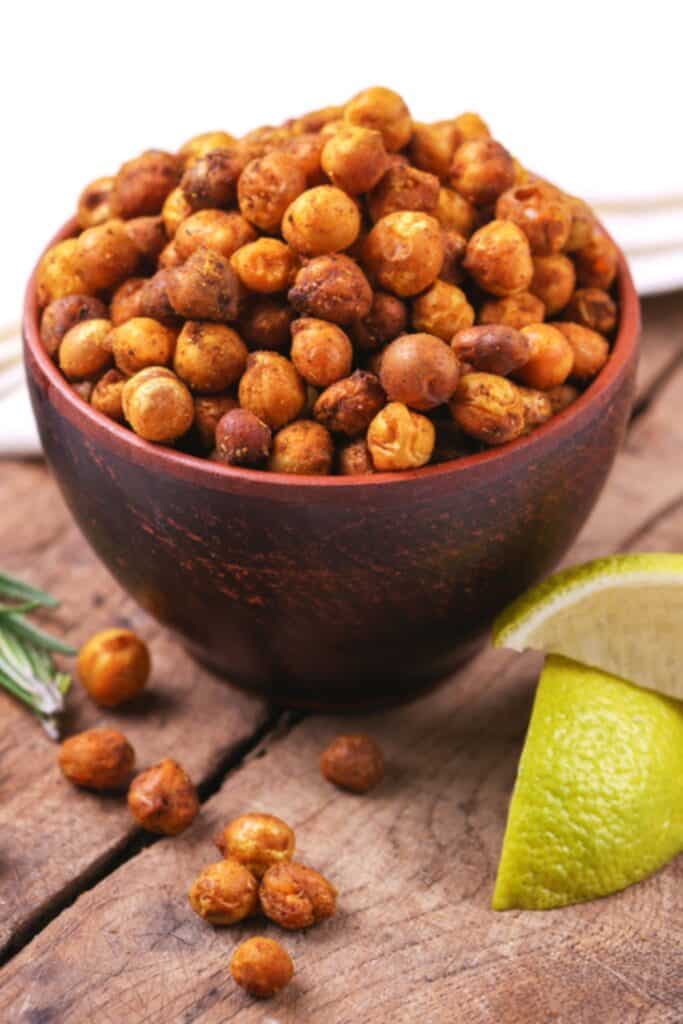 Close up of air fryer roasted chickpeas piled in a rustic wooden bowl, coated in golden brown seasoning with a crisp, slightly blistered texture. A few chickpeas are scattered on the wooden surface beside a fresh lime wedge and sprig of rosemary.