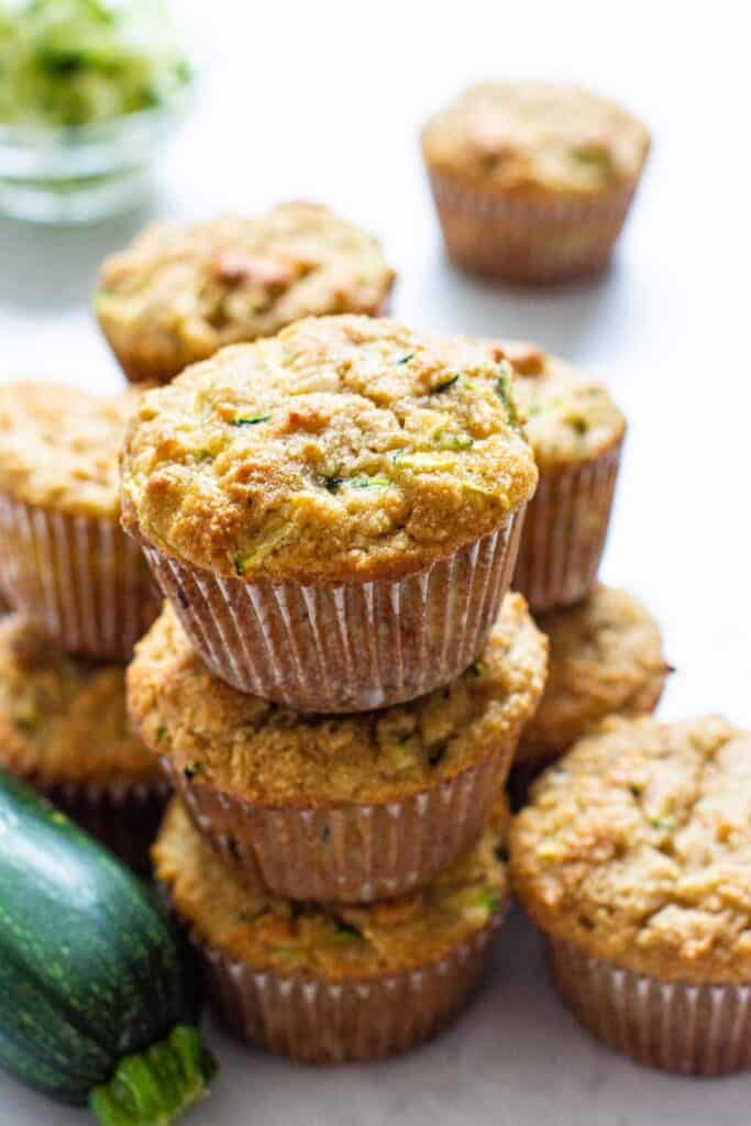 Stack of almond flour zucchini muffins with golden brown tops and a slightly crumbly texture, showing small green flecks of shredded zucchini throughout. The muffins sit in paper liners and are arranged in a tall stack, with a whole zucchini visible nearby.
