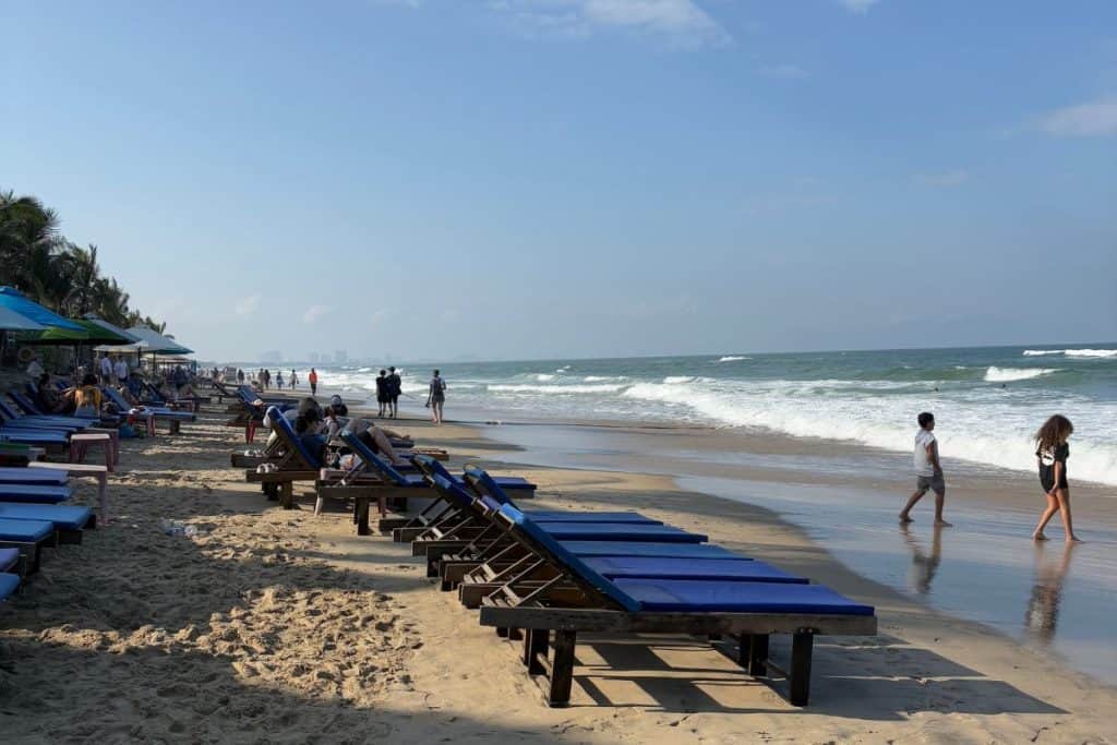 A row of wooden beach loungers with blue cushions lines a sandy shore under a sunny blue sky, with gentle waves rolling in and people walking along the water’s edge. The beach is lively but relaxed, with vacationers enjoying the coastal scenery.