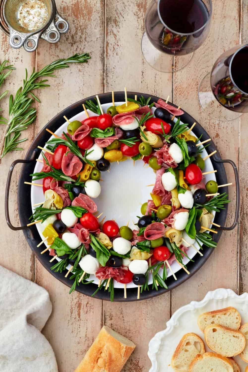 Antipasto charcuterie board arranged like a Christmas wreath with skewers of salami and prosciutto. Mozzarella balls cherry tomatoes green and black olives artichoke hearts peppers fresh basil and rosemary fill the wreath with sliced bread served alongside.