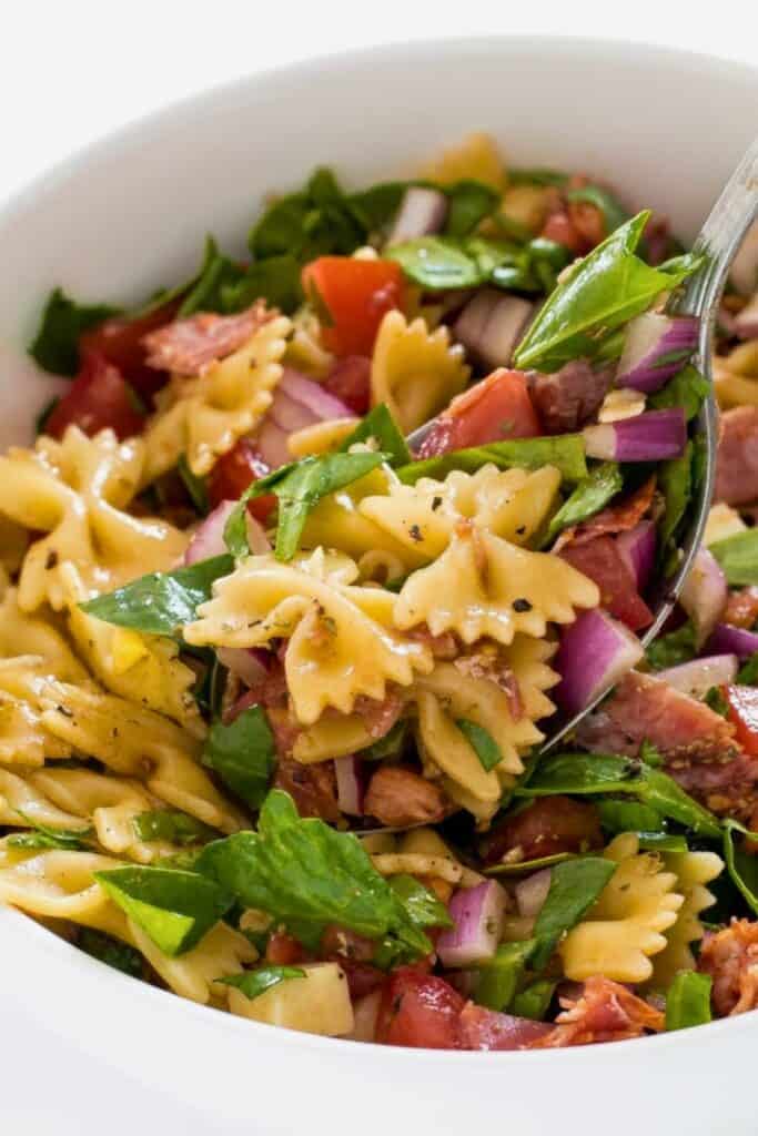 Bowtie pasta salad tossed with fresh spinach, diced red onion, cherry tomatoes, and chunks of salami in a light vinaigrette dressing. The colorful mix of vegetables and pasta creates a fresh and flavorful pasta salad.
