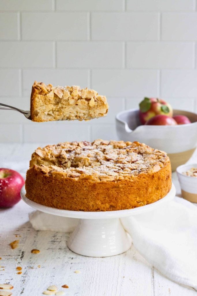 A golden apple almond cake sits on a white pedestal stand, with one slice being lifted to reveal its moist, apple-filled layers and crunchy almond topping. Lightly dusted with powdered sugar, the cake is surrounded by fresh apples and rustic decor.