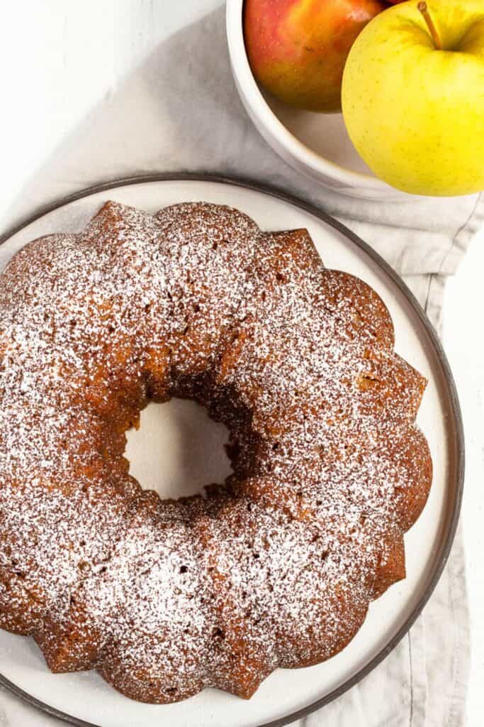 A golden brown apple bundt cake is dusted with powdered sugar and displayed on a round white plate, highlighting its beautifully scalloped edges.