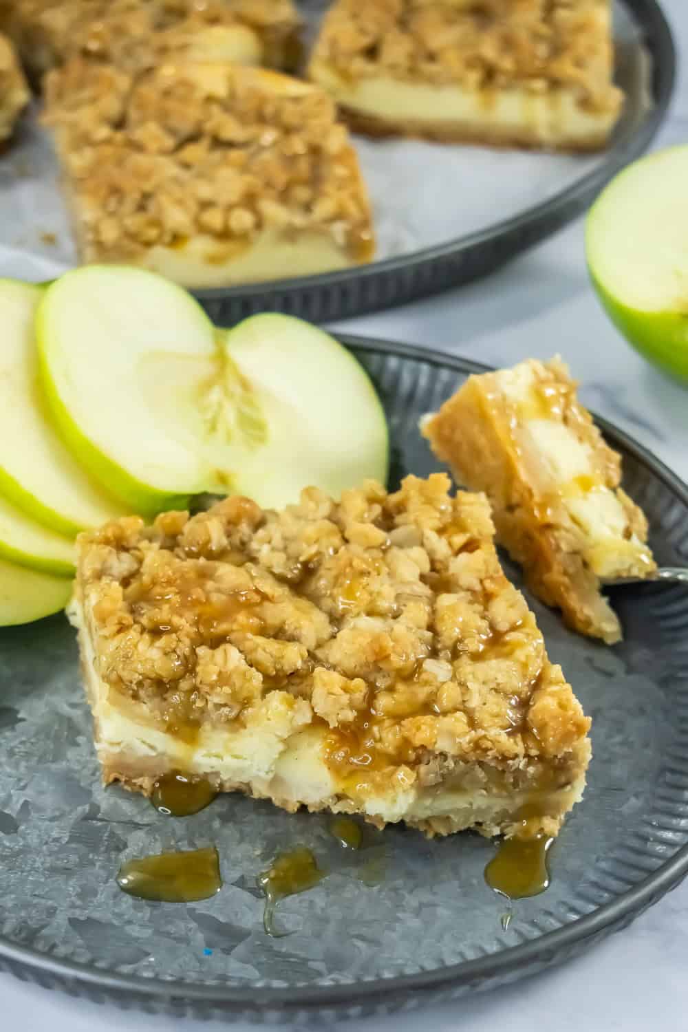 Caramel apple crisp cheesecake bars with a creamy cheesecake layer and crumbly oat topping, drizzled with caramel sauce and served with sliced green apples.