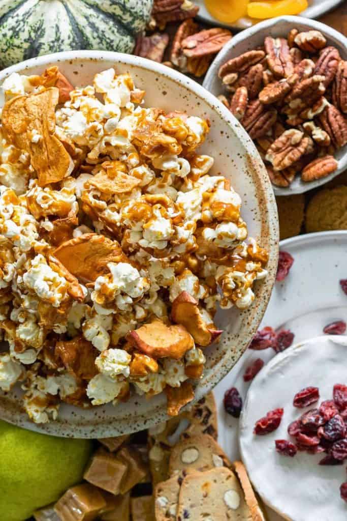 Close-up of a rustic bowl filled with apple caramel popcorn, featuring fluffy popcorn coated in golden caramel and mixed with dried apple slices. Surrounded by pecans, caramels, fruit, and cheese, this snack is part of a cozy, autumn-inspired grazing spread.
