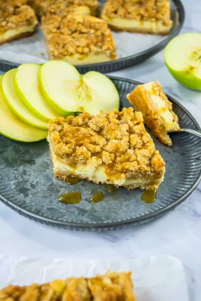 A caramel apple cheesecake bar sits on a metal plate next to thin slices of green apple, topped with a golden oat crumble and drizzled with rich caramel sauce. The layered dessert combines creamy cheesecake, tender apple chunks, and a buttery crust for a decadent fall treat.