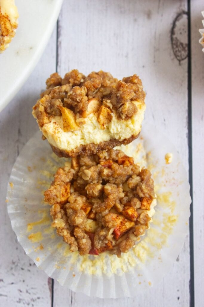 Two apple cheesecake bites topped with a golden brown cinnamon oat crumble sit on a white cupcake liner, one stacked atop the other to show the creamy cheesecake filling and graham cracker crust. The spiced apple chunks and crumbly topping make these festive mini desserts perfect for a Thanksgiving treat.