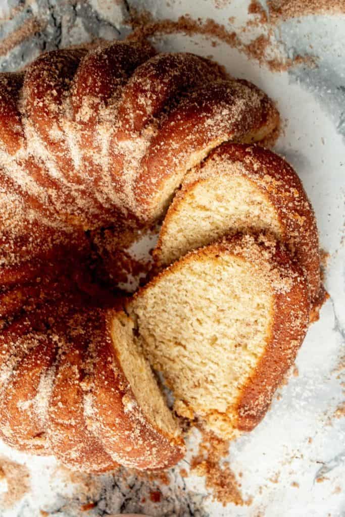 Close-up of a bundt-style apple cider donut cake coated in a generous layer of cinnamon sugar, with a few slices cut to reveal its soft, moist interior. The sugary topping mimics the texture of a classic donut.