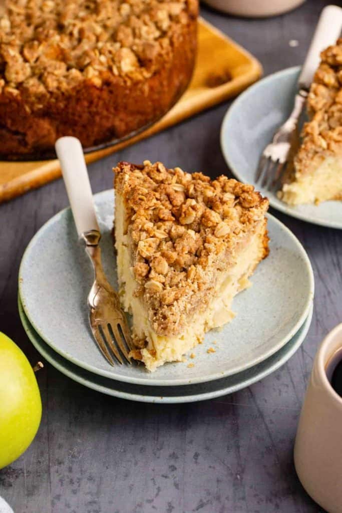 Slice of apple coffee cake topped with a golden oat streusel, served on a light blue plate with a fork. The creamy cheesecake and crumbly topping are paired with coffee and a green apple in the background. 