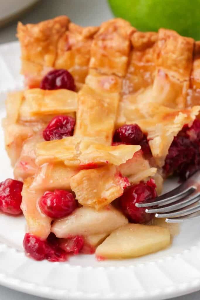 A slice of apple cranberry pie rests on a white plate, filled with tender apple slices and bright red cranberries, all tucked beneath a golden, flaky lattice crust.