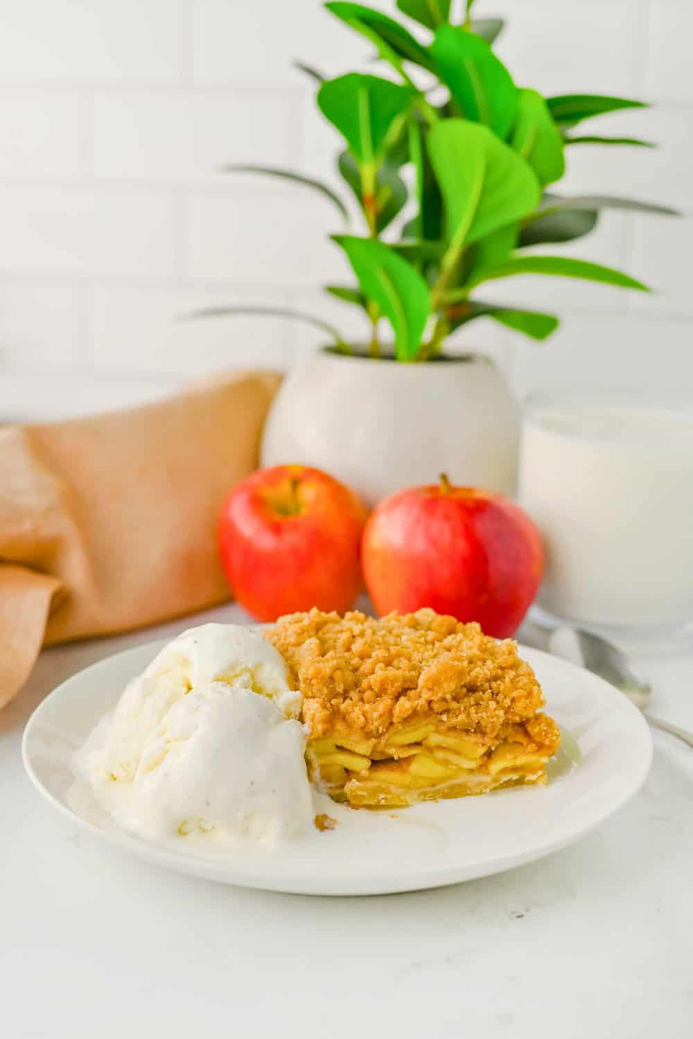Apple crisp bars layered with sliced cinnamon apples and a golden crumb topping served with melting vanilla ice cream. Two red apples and a green potted plant sit in the background.
