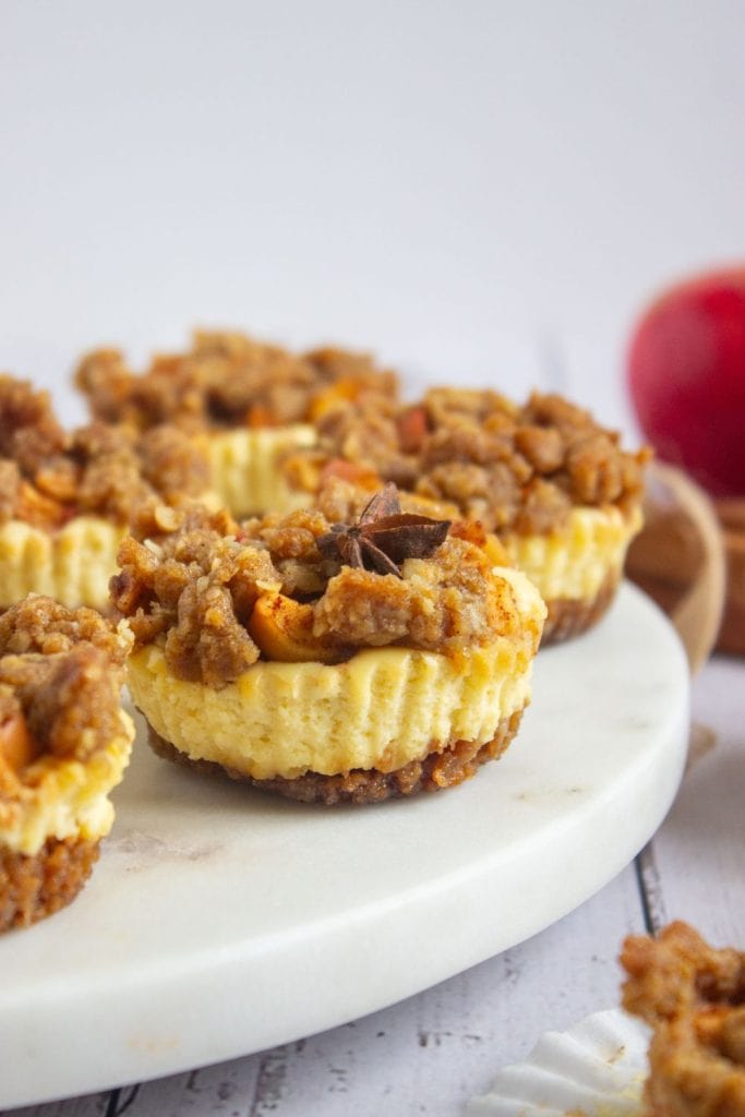 Close-up of a mini apple crumble cheesecake topped with spiced apples, oat streusel, and a decorative star anise. The cheesecake sits on a marble platter, with more mini cheesecakes blurred in the background.