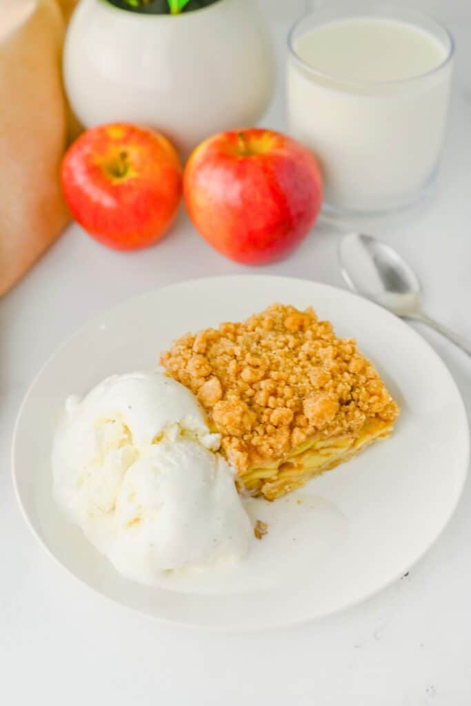 Alt text: Apple crisp bars with layers of cinnamon apples and a golden crumble topping served with melting vanilla ice cream. Two red apples and a glass of milk sit behind the plated dessert.