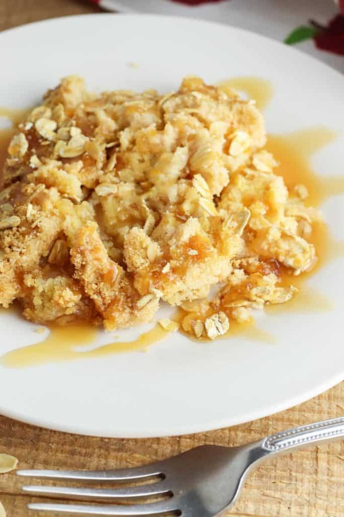 A serving of apple dump cake on a white plate, with tender, baked apple filling and a buttery, golden cake topping sprinkled with oats. Caramel sauce is drizzled over the top, enhancing the warm, rustic presentation.
