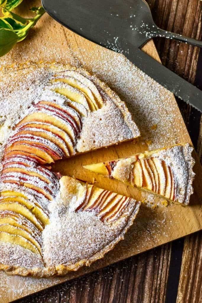 A rustic apple tart dusted with powdered sugar, featuring thinly sliced red and yellow apples arranged in a fan pattern. One slice is pulled out to show the tender, golden crust and soft baked filling, making it a perfect example of cozy fall baked goods.
