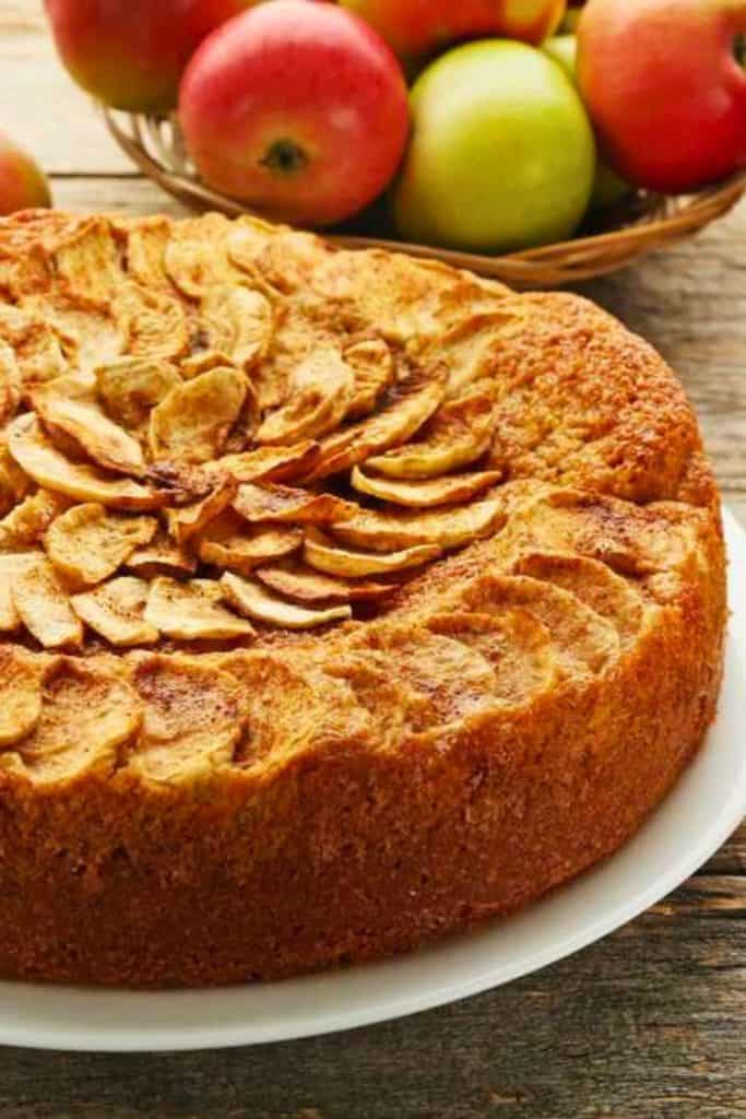 A rustic apple kuchen with a golden, cake-like base topped with spiraled slices of cinnamon-dusted apples, served on a white plate. A basket of red and green apples are in the background.