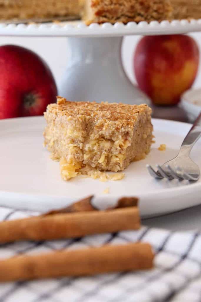 A close-up of a slice of moist apple cake with a crumbly, golden top, showcasing bits of grated apple and a soft, dense texture. The slice is served on a white plate beside a fork, whole apples and cinnamon sticks.