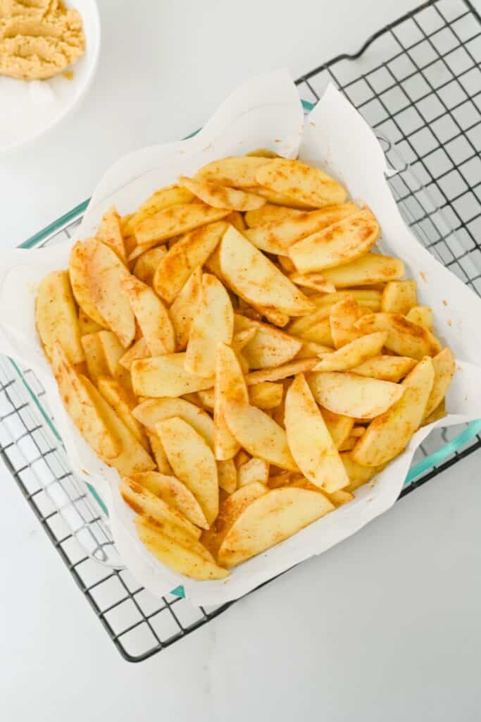 Cinnamon spiced apple slices spread over the crust in a parchment lined baking dish while assembling apple pie bars. The apples form a thick even layer ready for the crumb topping.